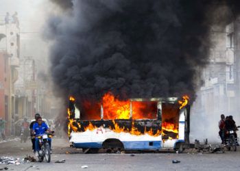 Dos motocicletas pasan junto a un autobús en llamas, incendiado por manifestantes de oposición que exigen saber el destino de los fondos de Petrocaribe, en Puerto Príncipe, Haití, el domingo 18 de noviembre de 2018. (AP Foto/Dieu Nalio Chery)