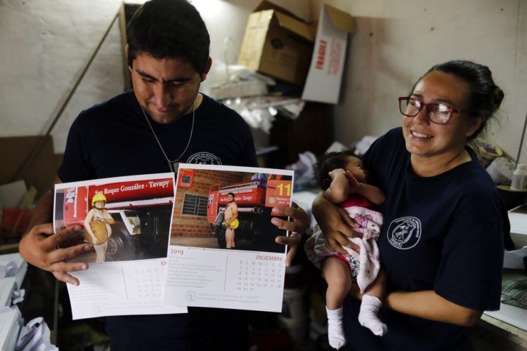 La bombera Fátima Olmedo (derecha) sostiene a su hija de dos meses Samara mientras su esposo, Rodrigo Giménez, muestra las páginas del calendario donde salen desnudos, en Asunción, Paraguay. Foto: Jorge Saenz / AP.