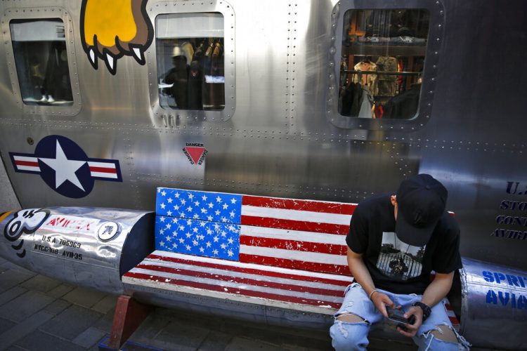 Un hombre navega en su smartphone en una banca decorada con la bandera estadounidense afuera de una tienda que vende marcas de Estados Unidos en un popular centro comercial en Beijing, el lunes 13 de mayo de 2019. (AP Foto/Andy Wong)