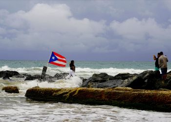 Una mujer posa para una fotografía con el mar y una bandera de Puerto Rico de fondo tras el paso de la tormenta tropical Dorian por la isla, en San Juan, Puerto Rico, el 28 de agosto de 2019. Foto: Ramón Espinosa/ AP.