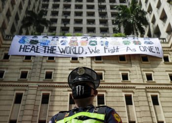 Un guardia de seguridad de pie ante una pancarta que dice "Sanen al mundo, luchamos como uno", mientras los vecinos aplauden desde las ventanas en homenaje a sanitarios, personal esencial y fuerzas de seguridad, durante una cuarentena impuesta para impedir la expansión del nuevo coronavirus en Manila, Filipinas, el domingo 12 de abril de 2020. (AP  Foto/Aaron Favila)