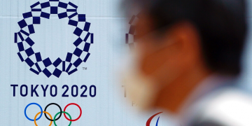 Un hombre con una mascarilla protectora contra infecciones respiratorias como la COVID-19 pasa junto al logo de los juegos olímpicos Tokio 2020. Foto: Eugene Hoshiko / AP / Archivo.