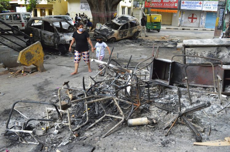 Una mujer y un niño pasan junto a los restos de vehículos y muebles quemados en las protestas de Bengaluru, India, el miércoles 12 de agosto de 2020. Al menos tres personas murieron en la ciudad en choques entre la policía y manifestantes que protestaban por una publicación de Facebook que consideraban como una ofensa al islam. Foto: AP.