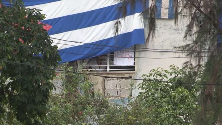 El activista Yunior García Aguilera, uno de los convocantes de la marcha del 15 de noviembre, se asoma por su ventana con flores en la mano, mientras agentes vestidos de civil cubren su ventana con una bandera cubana. Foto tomada de la cuenta en Twitter del periodista Atahualpa Amerise.