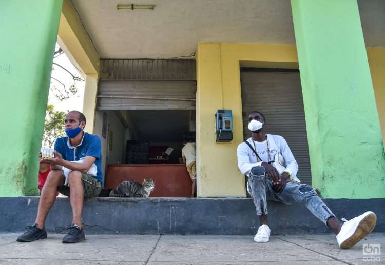 Hombres sentados delante de una bodega, uno de ellos sostiene varias cajetillas de cigarro. Foto: Kaloian.