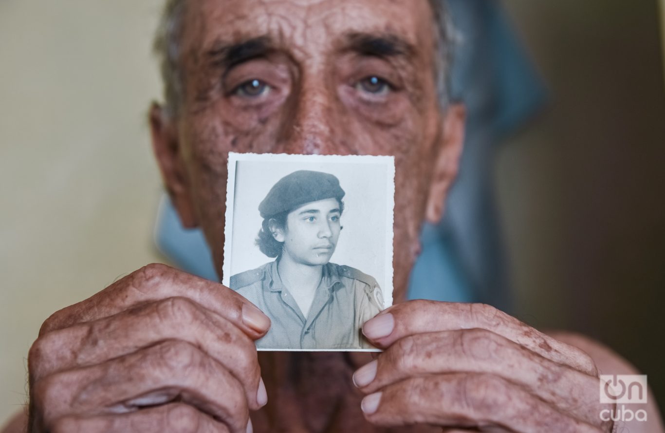 Delfín posa sosteniendo una fotografía de sí mismo de cuando era joven, en 1961, durante su época como alfabetizador en la Sierra Maestra. Foto: Kaloian.