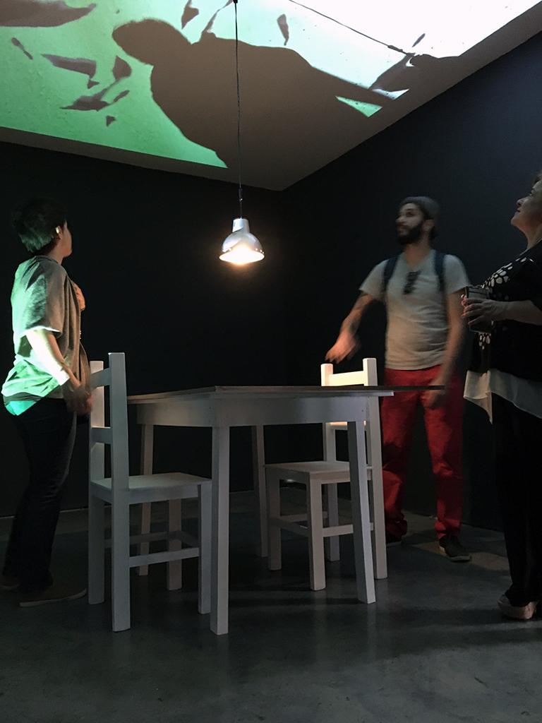 Detalle de la instalación “Emboscada–Habeas Paraíso”. Bienal de Curitiba, Brasil, 2017. Público interactuando con la pieza, concebida como un set de interrogatorio. En el techo, proyección de video compuesto con imágenes de archivo y canales públicos. Foto: cortesía del artista.
