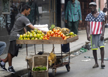 Un hombre con camiseta con la bandera de Estados Unidos pasa frente a un vendedor ambulante de verduras, en La Habana. Foto:  Yander Zamora/EFE.